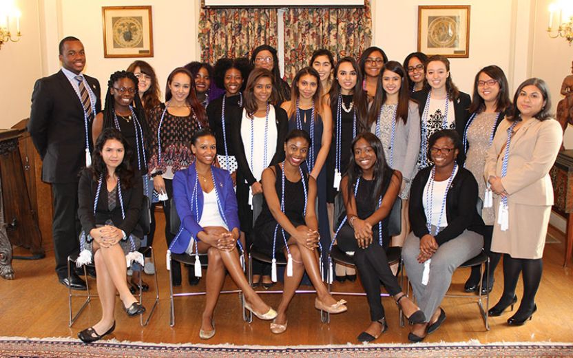 large group of students and staff dressed in professional attire wearing blue and white chords standing and sitting in a great hall at the AD White House on Cornell’s campus