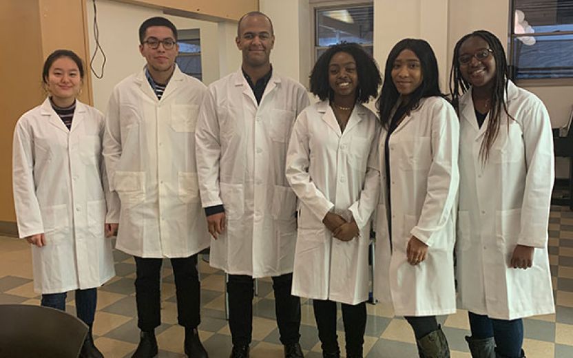 A group of six students in their white doctor's coats standing in a lab setting.]