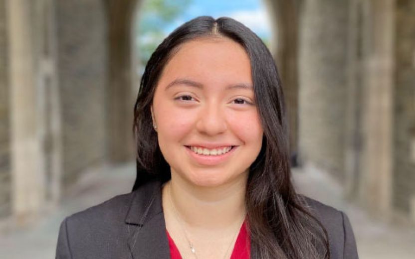 Photo of Yesenia in a black suit with red shirt standing in the arches of the gothic buildings on Cornell University's west campus housing area,