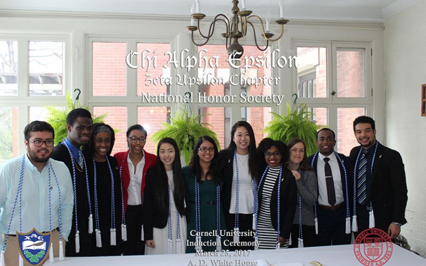 large group of students dressed in professional attire wearing blue and white chords standing in a white room surrounded by windows and a chandelier over head at the AD White House on Cornell’s campus