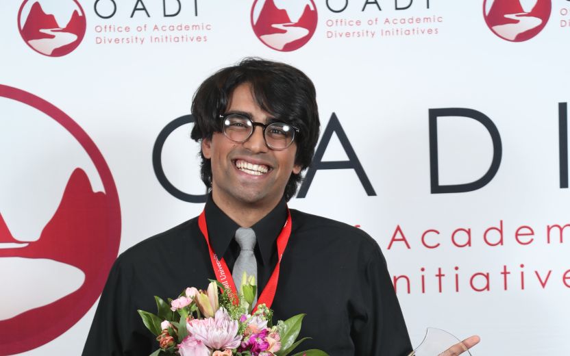 Adam is smiling as he is standing in from of a OADI backdrop and has a bouquet of flowers and a award in his hands