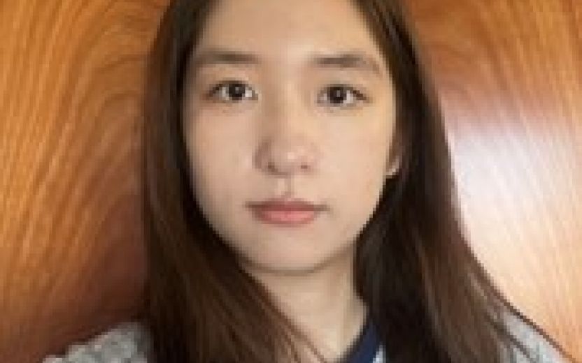 Yi Lin is looking at the camera and standing in front of a wood wall