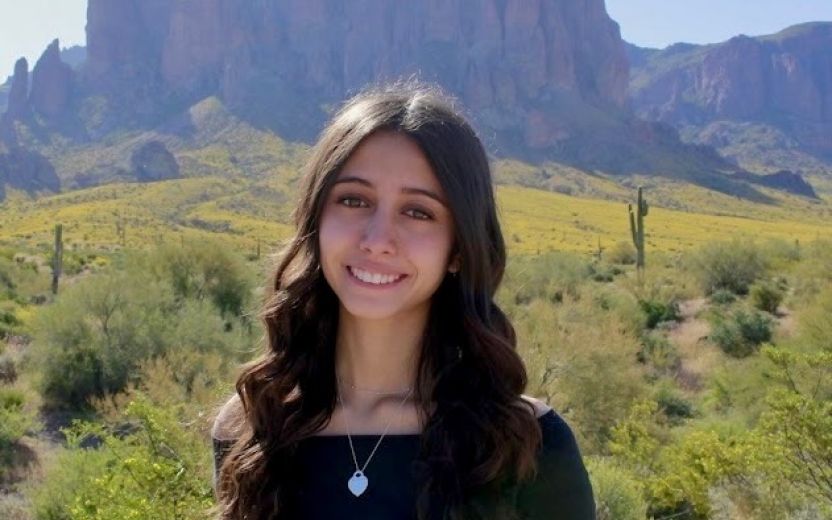 Allie Pequeno is standing somewhere outside in a desert landscape. She is smiling at the camera