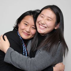 Two students hug each other and smile. One of them has a blue and white cord draped around the next