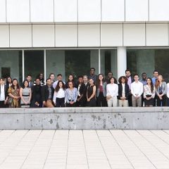 A group of around 50 students stand together to pose for a photo in a concrete courtyard outside of a white building.