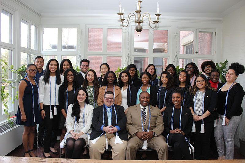 large group of students and staff dressed in professional attire wearing blue and white chords standing and sitting in a white room surrounded by windows and a chandelier over head at the AD White House on Cornell’s campus