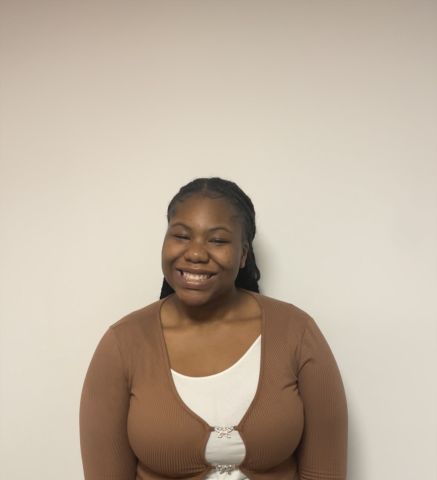 Ruth Osagie is smiling at the camera.  She is standing in front of a beige wall. She has on a light brown sweater over a white shirt.