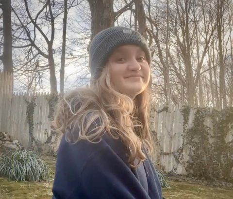 Sophie is smiling at the camera.  She is outside and has a gray winter hat and blue sweatshirt on