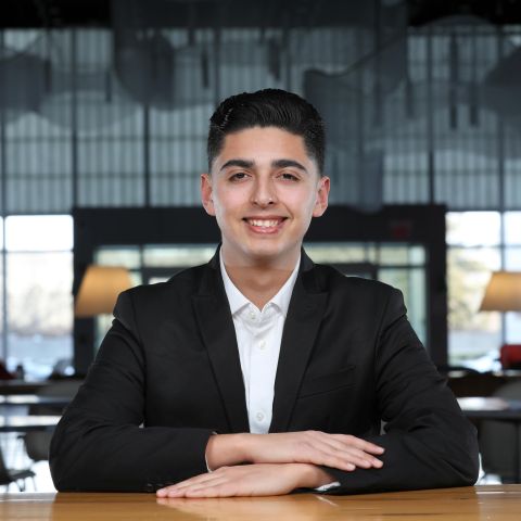 Diego Estrada is sitting down with his arms folded and smiling. He has on a blazer and button down shirt