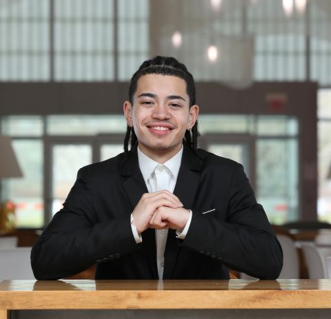 Shaunjae Suarez is sitting down and smiling at the camera. He has on a business suit