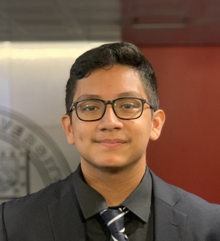 Headshot photo of Alain standing in front of a red wall in a black suit, black shirt, black and white tie.