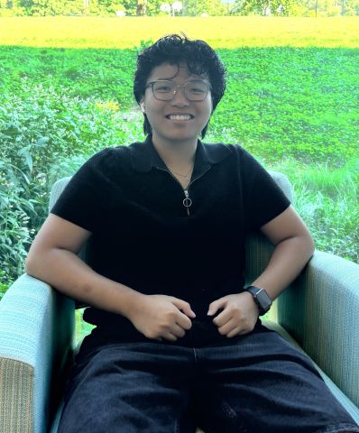 Photo of Michelle sitting in a chair in a black shirt and shorts in front of a tall window with grass and a tree behind her.