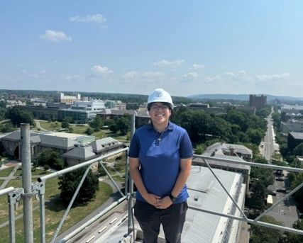  Teresa Crisantos-Prudencio is standing on a rooftop at Cornell with a hard hat on smiling at the camera