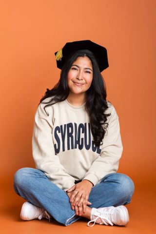 Cassie Guzman is sitting and smiling at camera, she has on a Syracuse shirt and a graduation cap