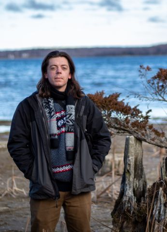 Christopher is standing outside at a beach and looking at the camera