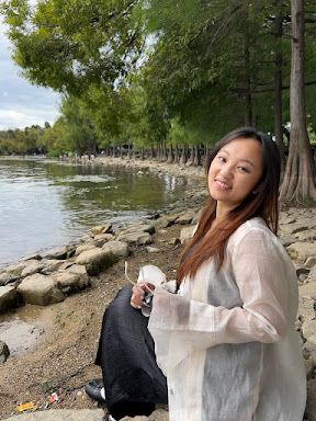 Ruier Dai is smiling at the camera. She is outside along the shore of a body of water.